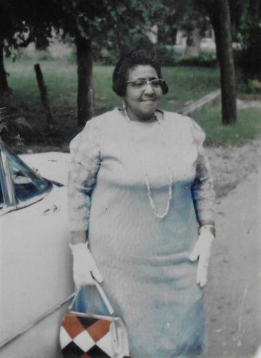 Woman in a light dress standing beside a car on a tree-lined path, holding a patterned handbag.