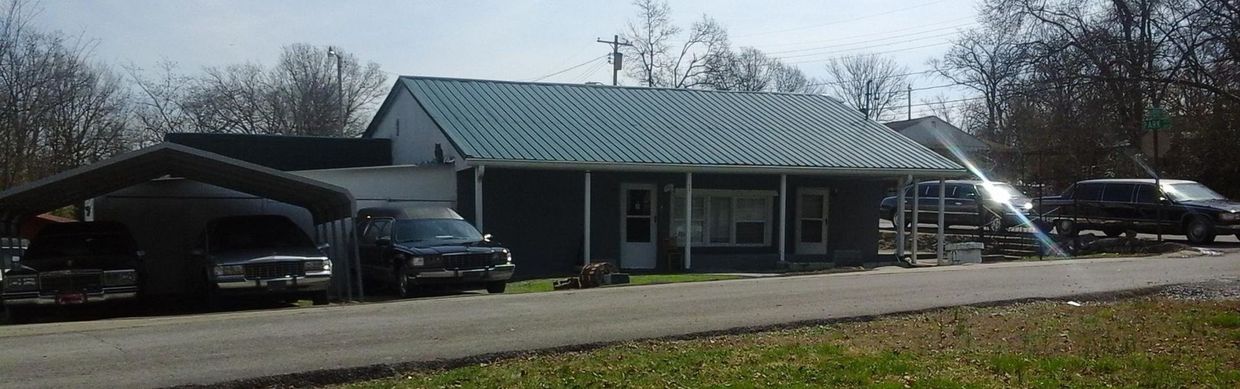 Small roadside building with a green metal roof and parked cars under a canopy, in a rural setting.