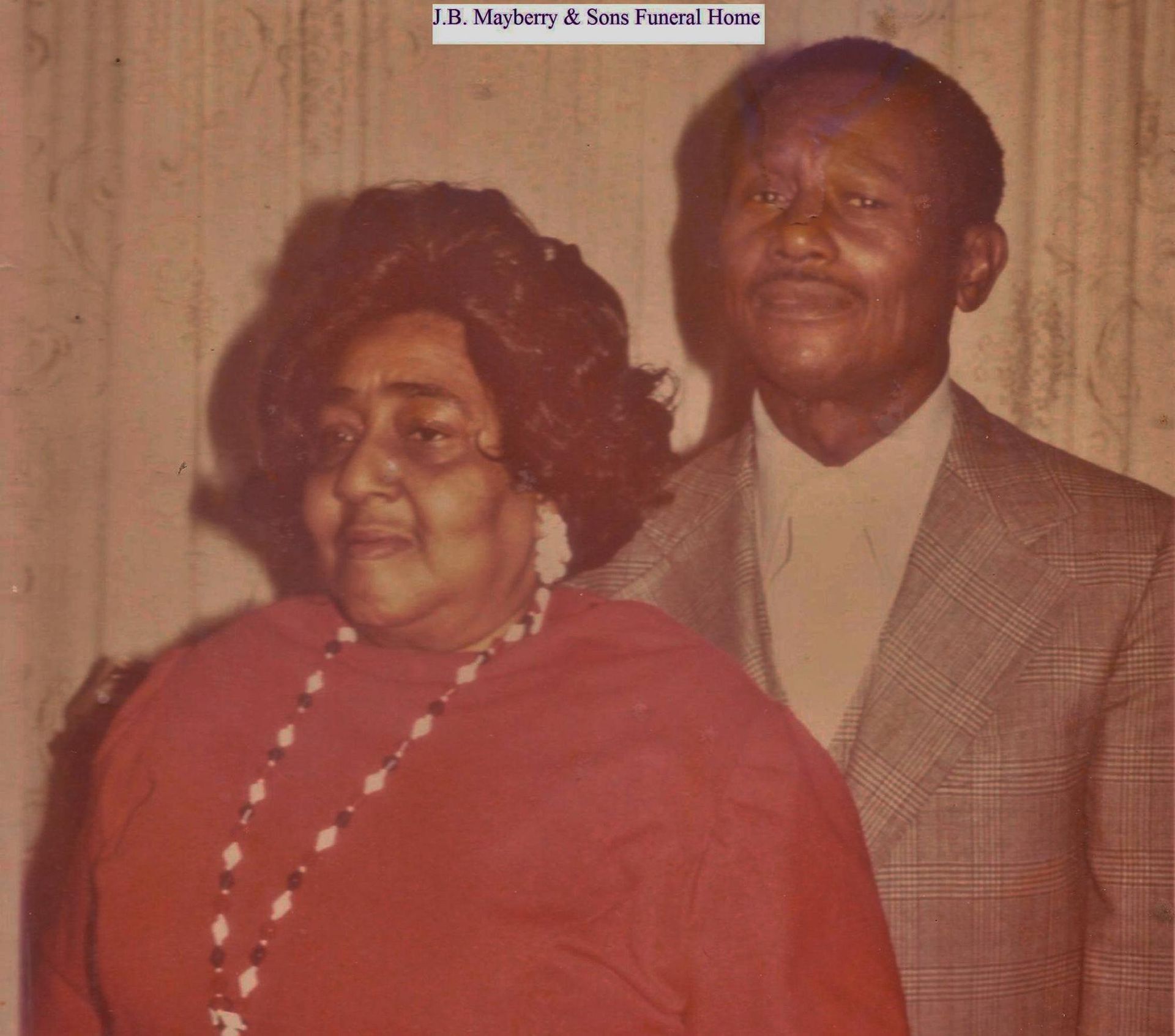 Two adults posing indoors; woman in red dress, man in brown suit, both facing the camera.