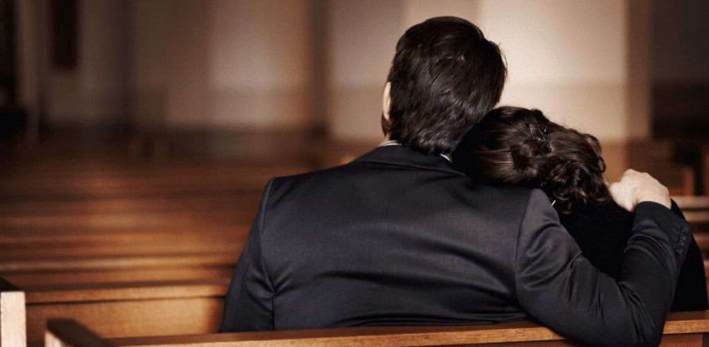 Two people sit together on church pews, one resting their head on the other's shoulder.