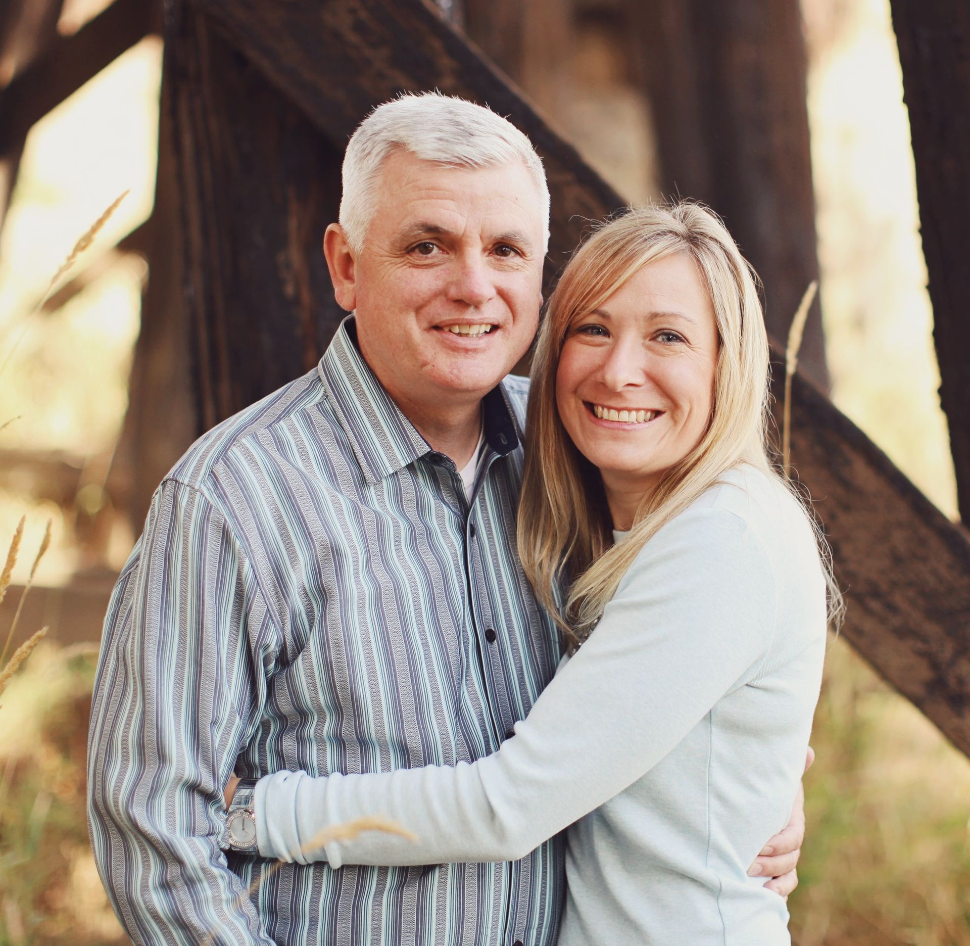 Smiling couple embracing outdoors near a wooden structure, with soft sunlight and blurred background.