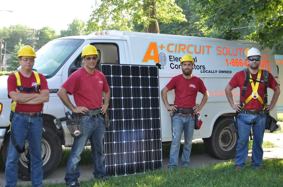Four workers in red shirts and hard hats stand with a solar panel in front of a white van with 