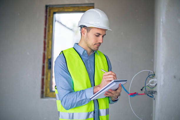 Construction worker in white hard hat and neon vest, writing on a notepad near electrical wiring.