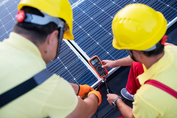 Two workers in yellow hard hats testing solar panels with a multimeter.