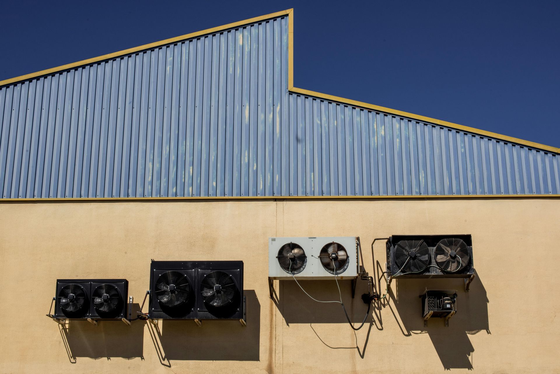 Industrial building facade with blue metal roof and beige wall, lined with mounted air conditioning units.