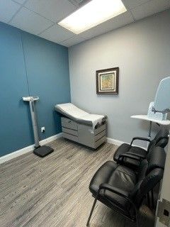 Medical exam room with exam table, scale, and two black chairs in a blue and gray interior.