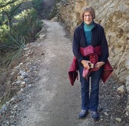 Person standing on a dirt trail beside rocky hillside, wearing a dark jacket and red sweater, smiling.