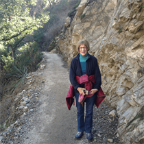 Person standing on a narrow mountain trail beside a rocky cliff, wearing a jacket and backpack straps.