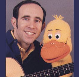 Man smiling beside a yellow puppet holding a guitar against a gray background