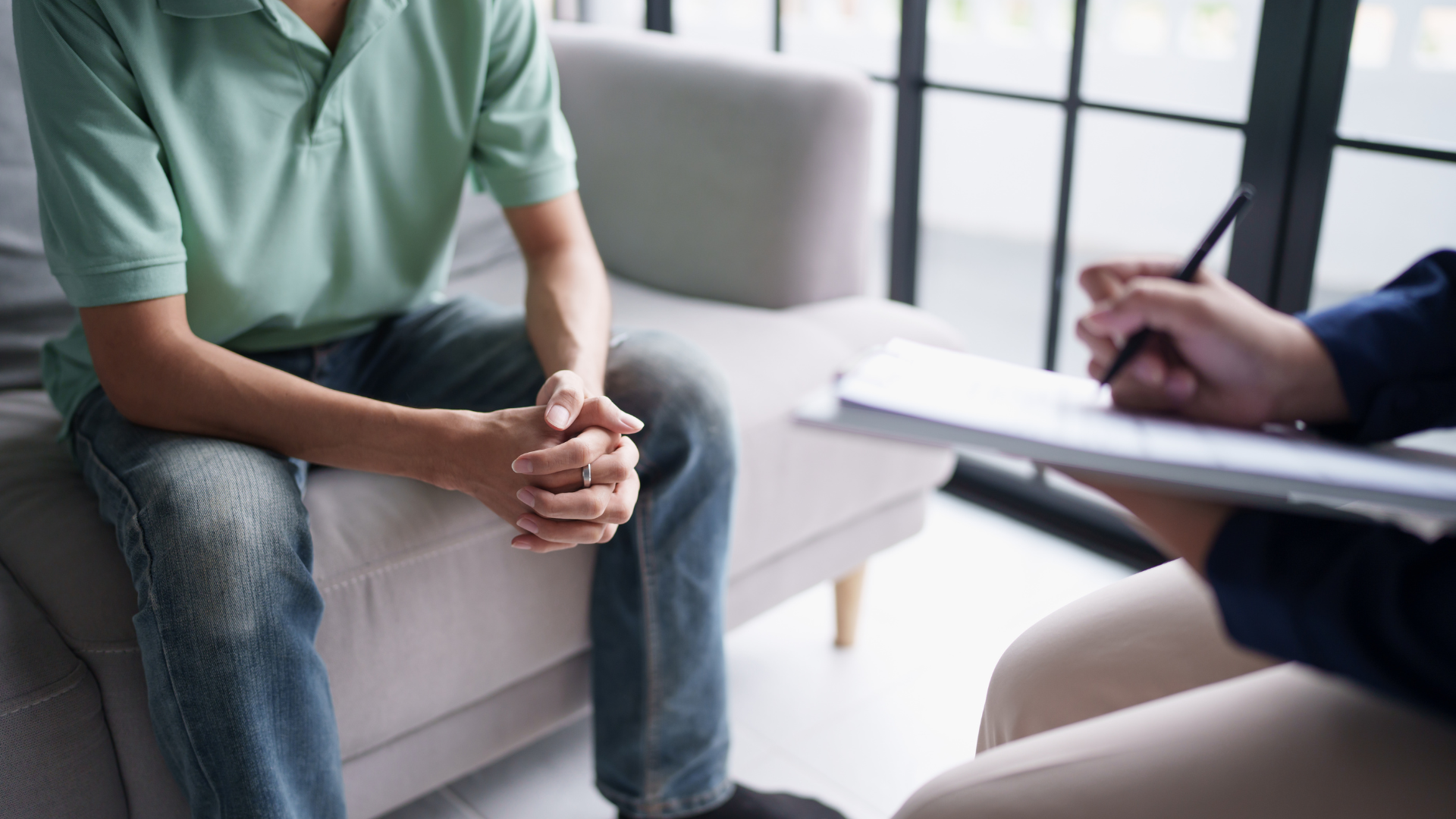 Man in therapy session; sits on sofa, hands clasped. Therapist takes notes on a clipboard.
