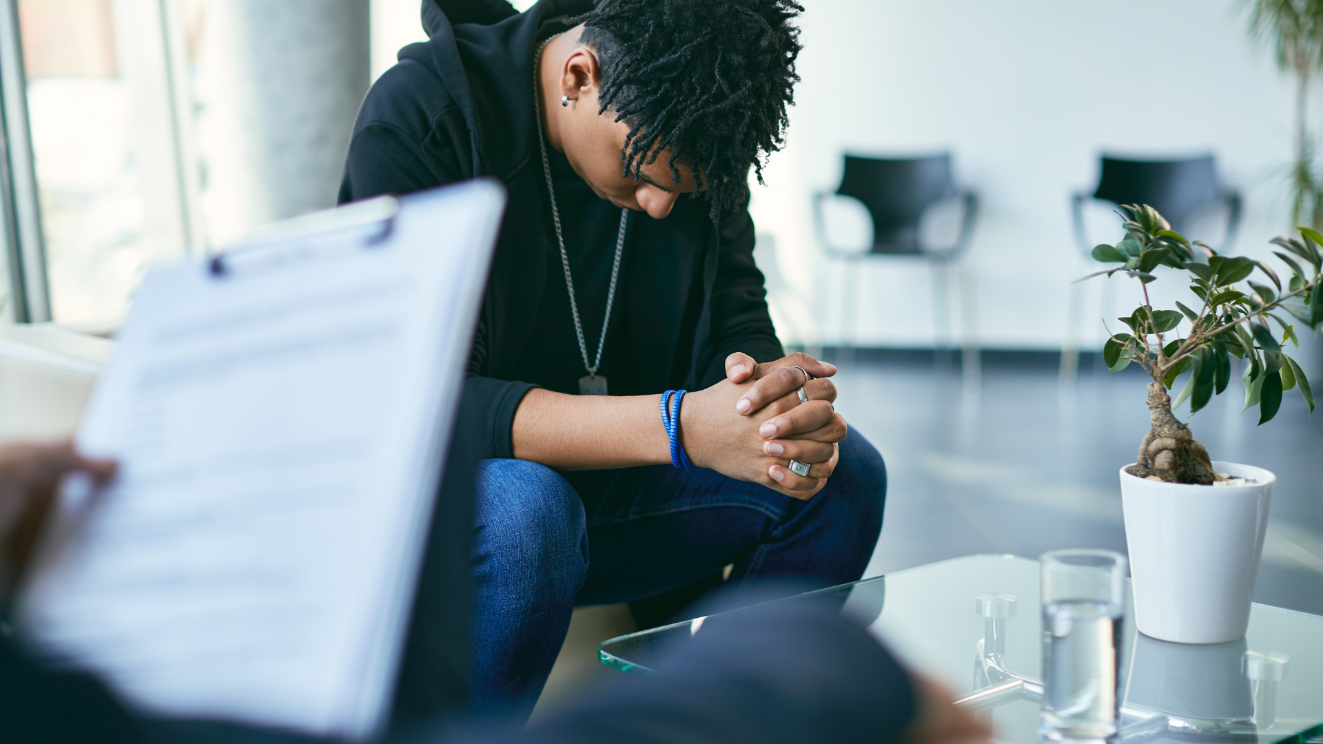 Person in therapy session, head bowed, hands clasped, listening to a clinician with a clipboard.