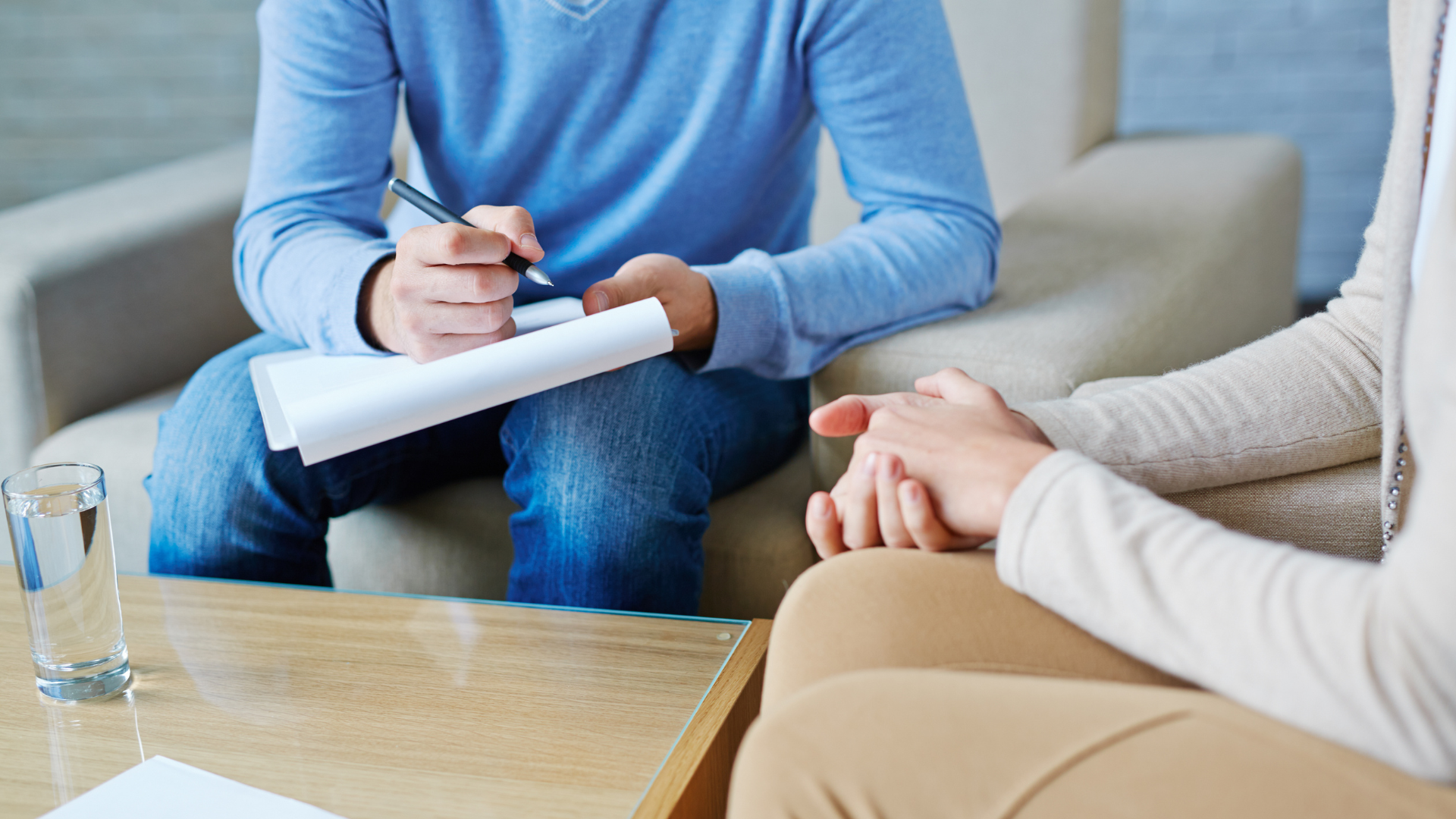 Person writing on paper, sitting with another person who has hands clasped, perhaps in therapy.