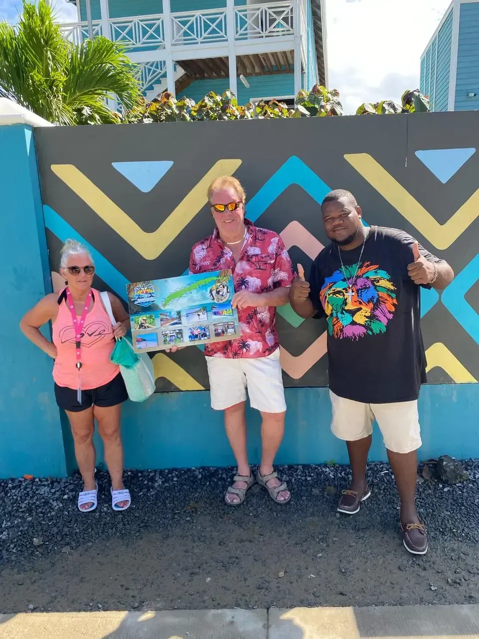 Three people stand by a colorful wall. Man holds photo display. One person gives thumbs up. Sunny outdoor setting.