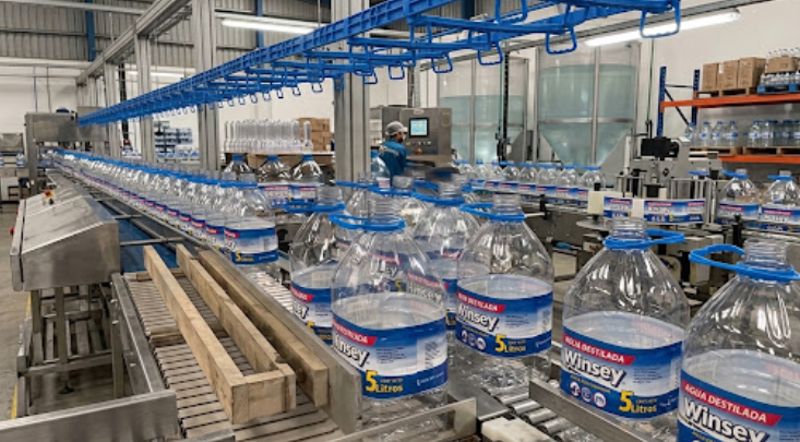 Filas de botellas de agua transparente con etiquetas azules y blancas sobre una mesa en una habitación luminosa.