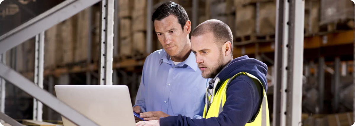 Worker in high-vis vest and leader in business shirt look at computer screen in warehouse.