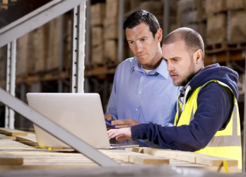 Two workers reviewing plans on a laptop in a warehouse, one in a yellow safety vest
