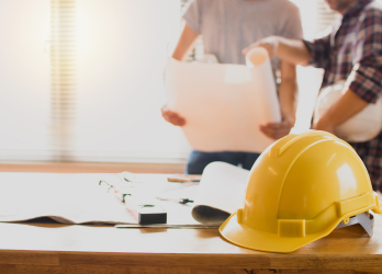 Yellow hard hat on a desk with people reviewing plans in the background