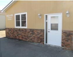Small tan building with brick lower wall, white door and window, and a paved driveway.
