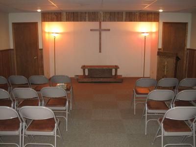 Small chapel with rows of folding chairs, wooden altar, and a cross on the front wall.