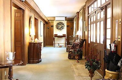 A wood-paneled hallway with a carpeted floor, lamps, a floral wreath, and a patterned armchair leading to double doors.