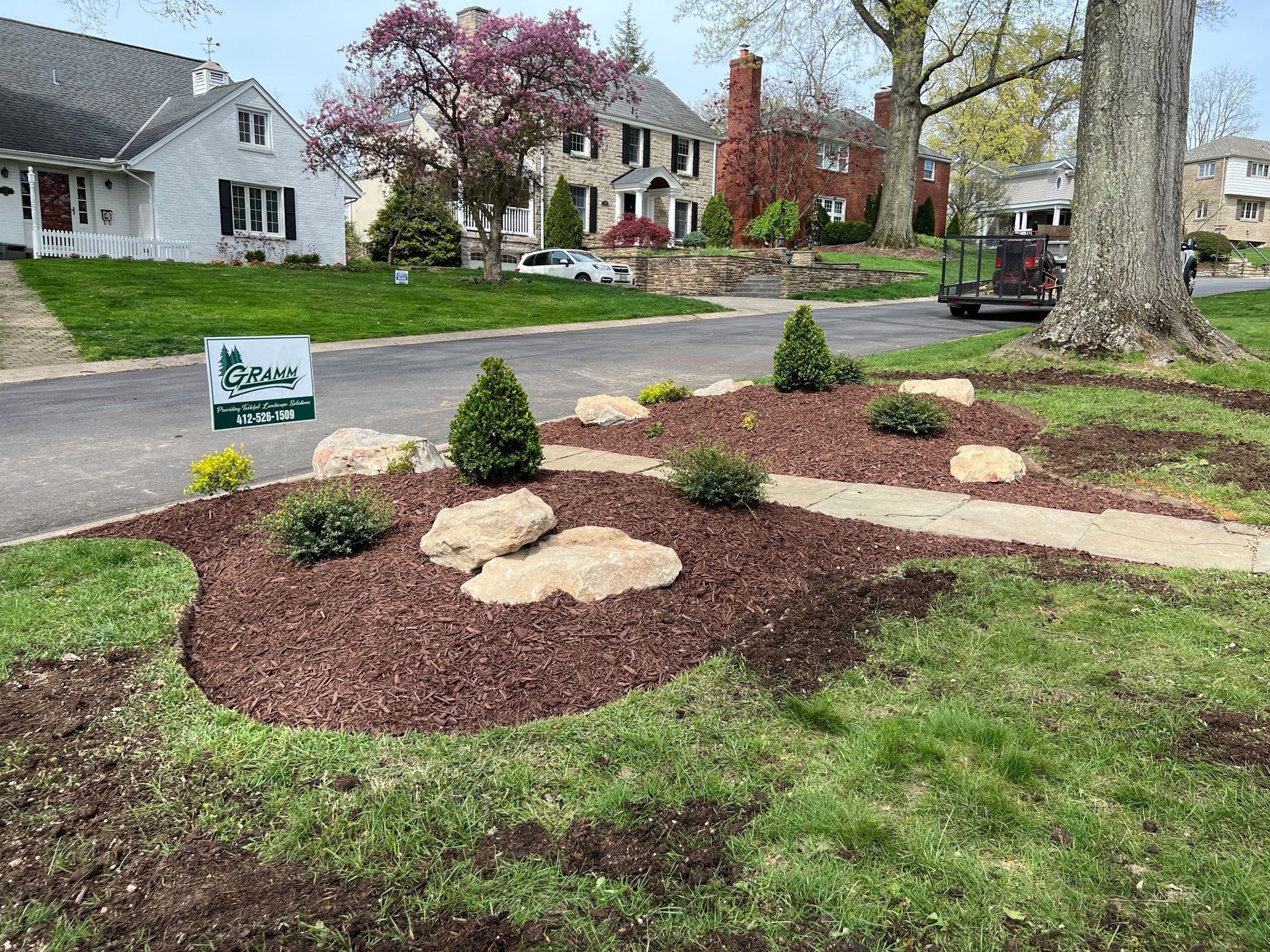 Landscaped front yard with mulch beds, shrubs, stepping stones, and a real estate sign on a suburban street