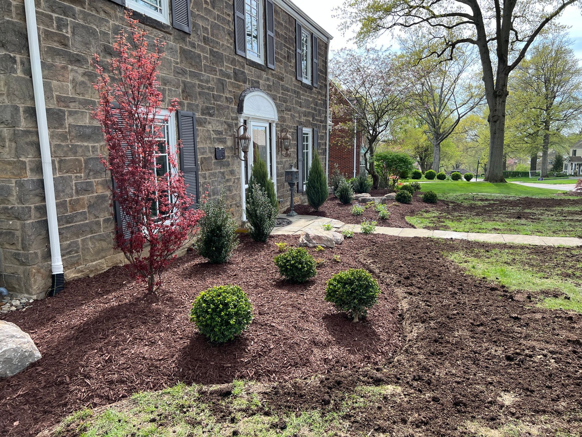 Landscaped yard beside a stone house, with mulch beds, shrubs, and a red-leaf tree.