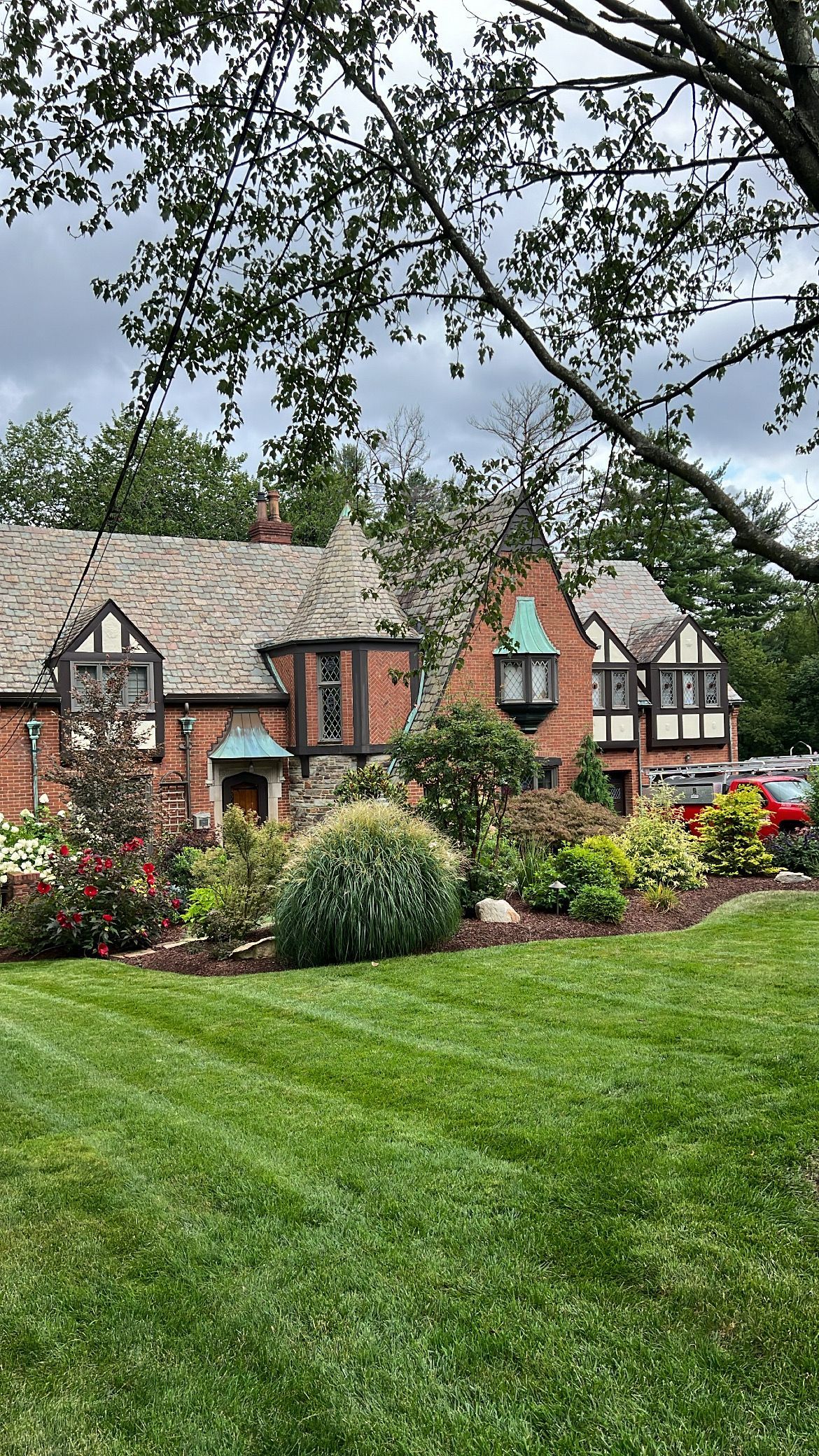 Brick house with gray roof, colorful flower garden, and a green lawn under tree branches