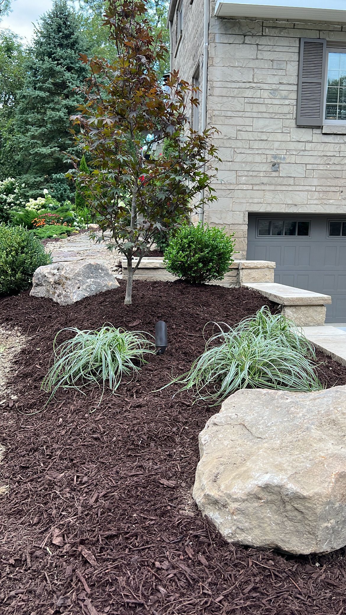 Landscaped front yard with mulch, rocks, shrubs, and a small tree beside a stone house exterior