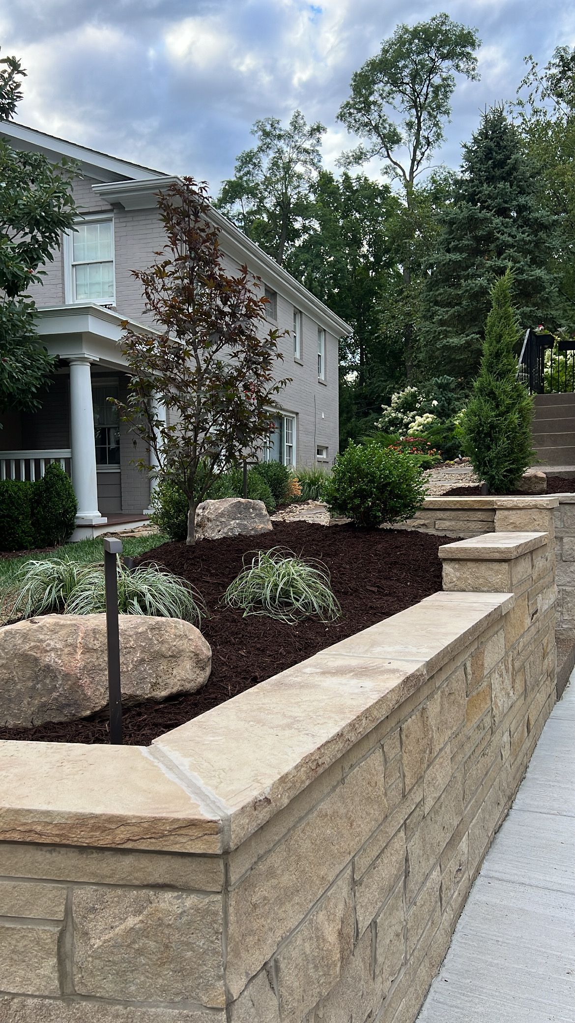 Stone retaining wall with landscaped shrubs beside a white house and sidewalk under cloudy sky