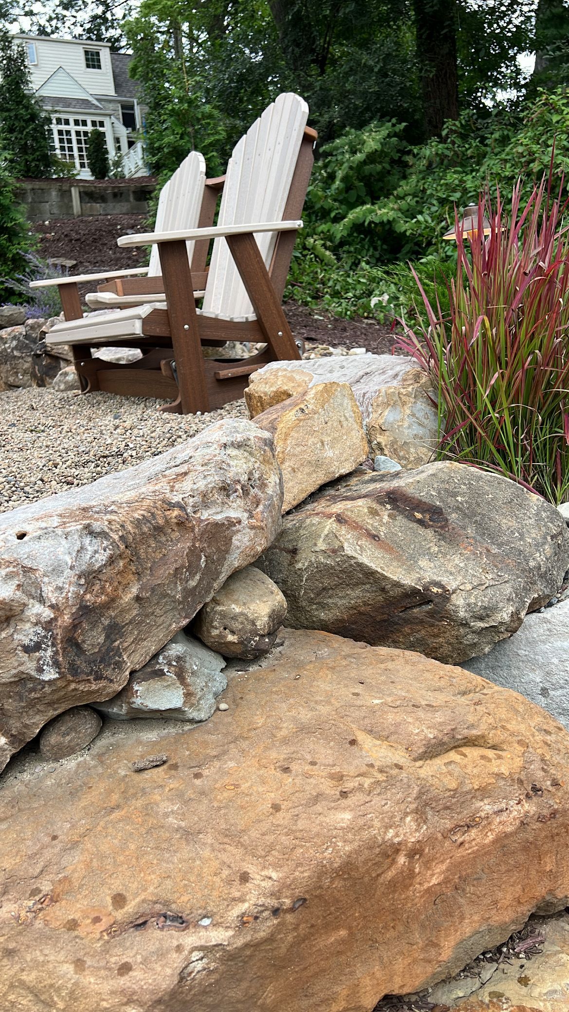 Two wooden Adirondack chairs on a rocky garden ledge beside flowers and a small stream