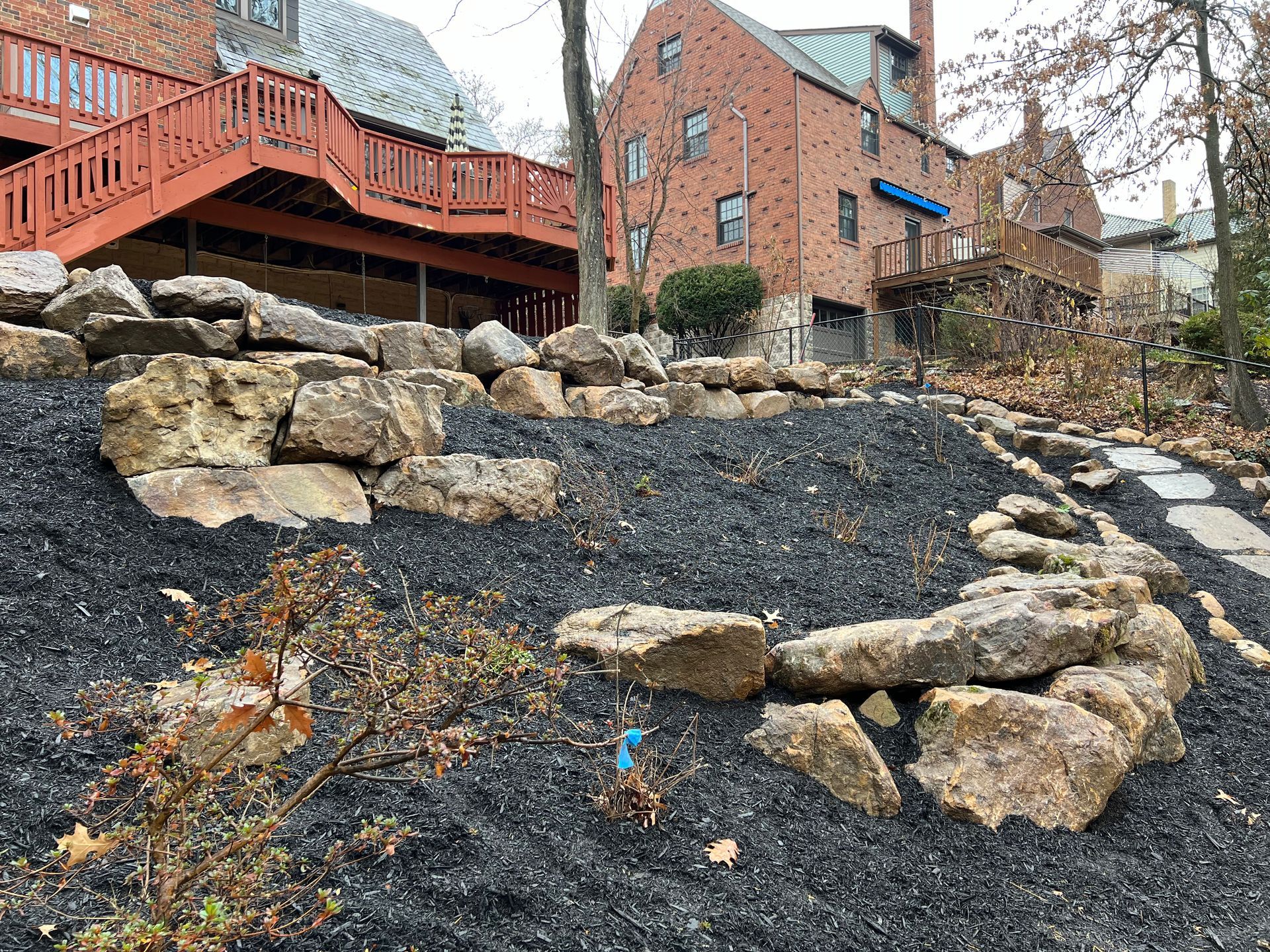 Stone retaining wall and black gravel landscaping below a brick house with a red wooden deck.