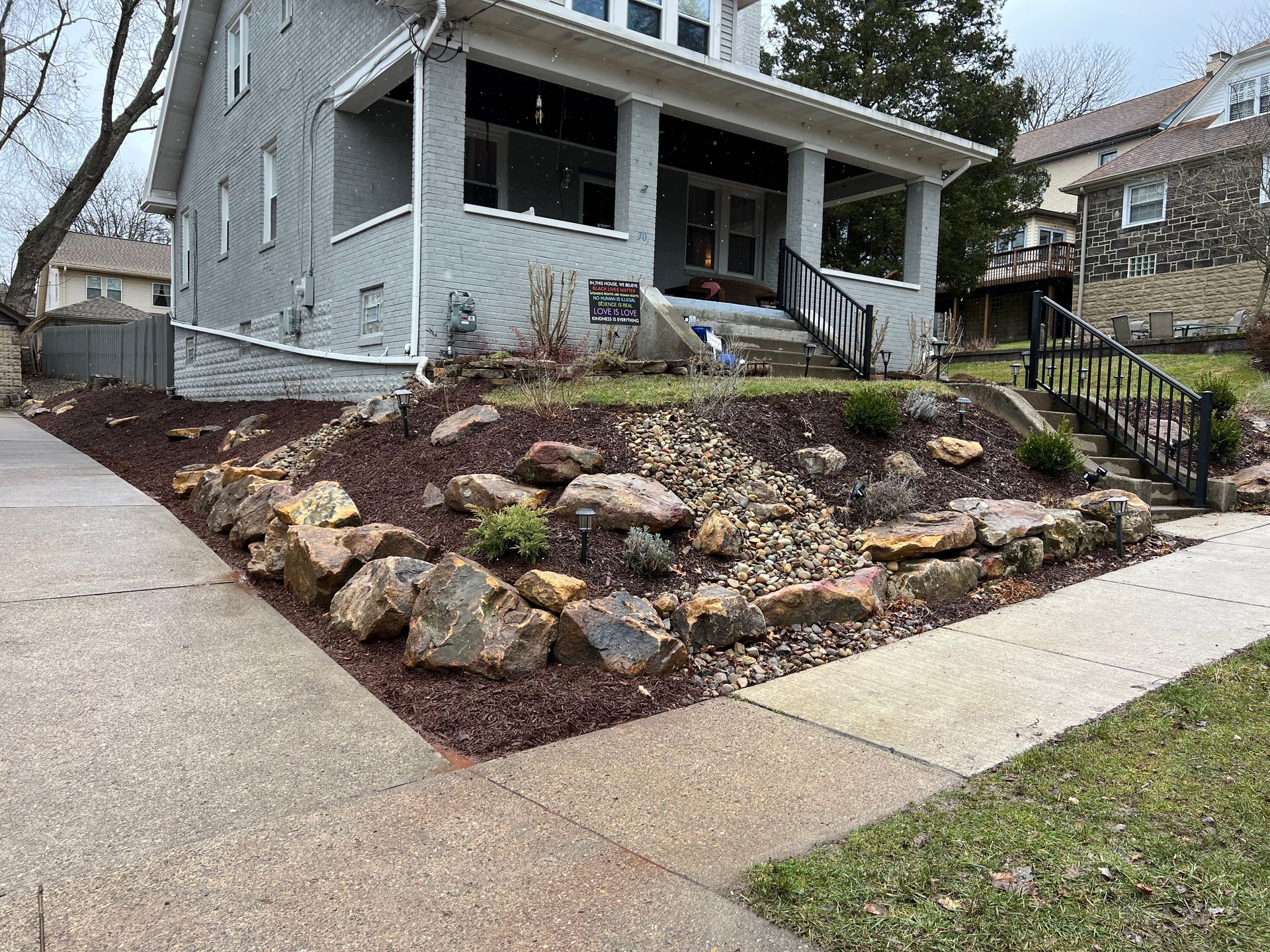 Gray house with a front porch and rock-covered sloped yard beside a sidewalk.