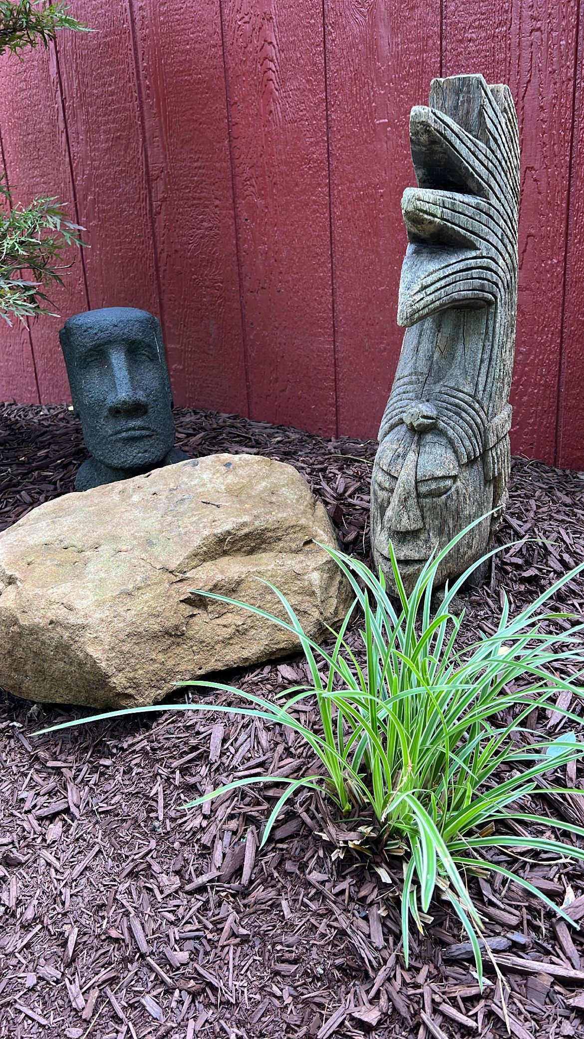 Two carved stone statues beside a large rock and green plant in a mulched garden against a red fence