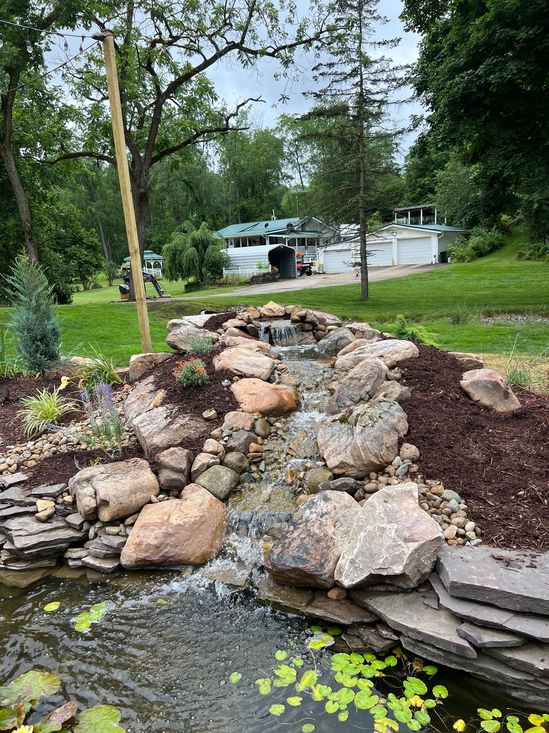 Rock garden with large stones, mulch, and a small waterfall in a leafy yard near a pool