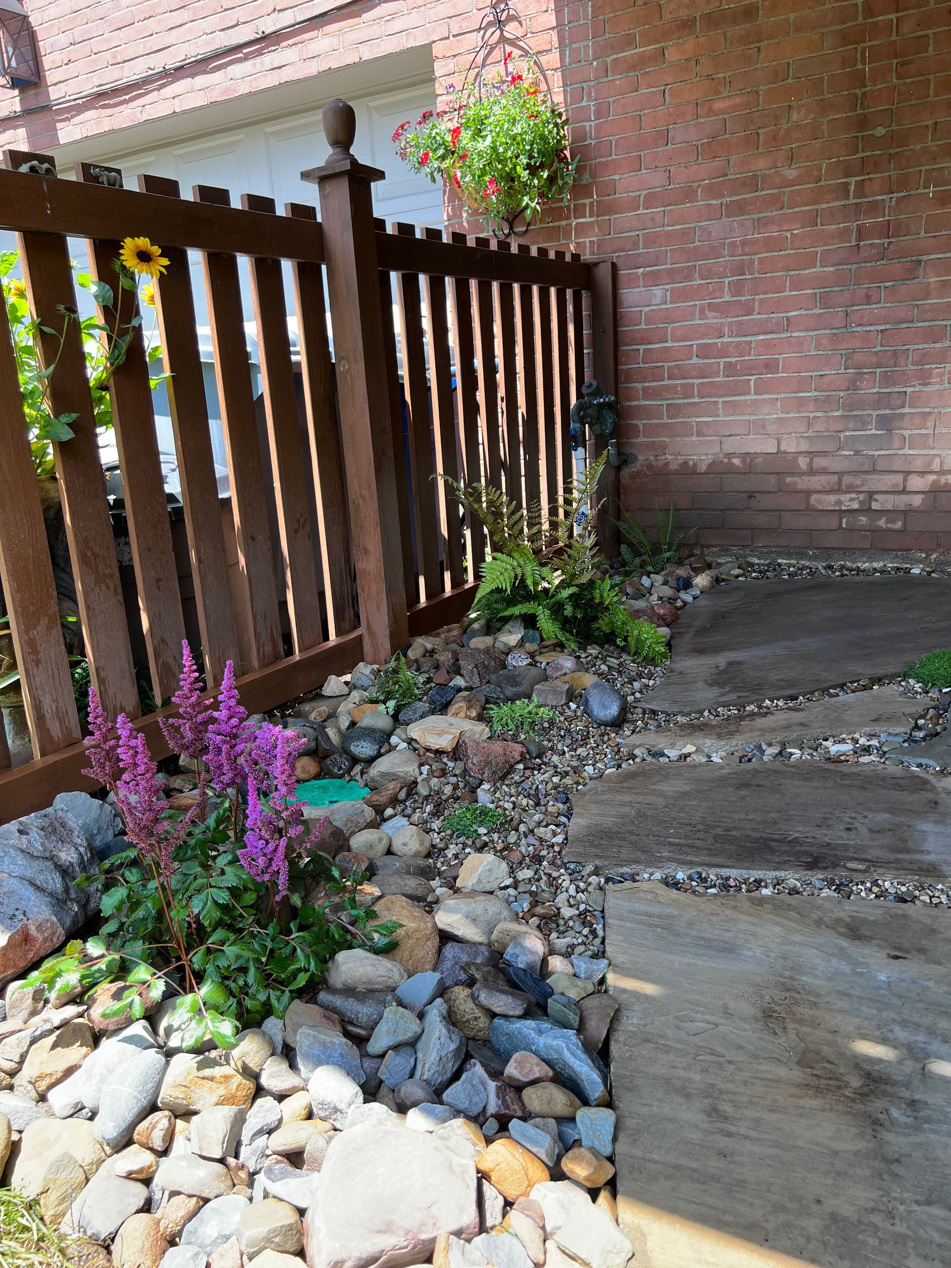 Brick patio with wooden fence, potted flowers, and a rock-bordered garden bed along a walkway
