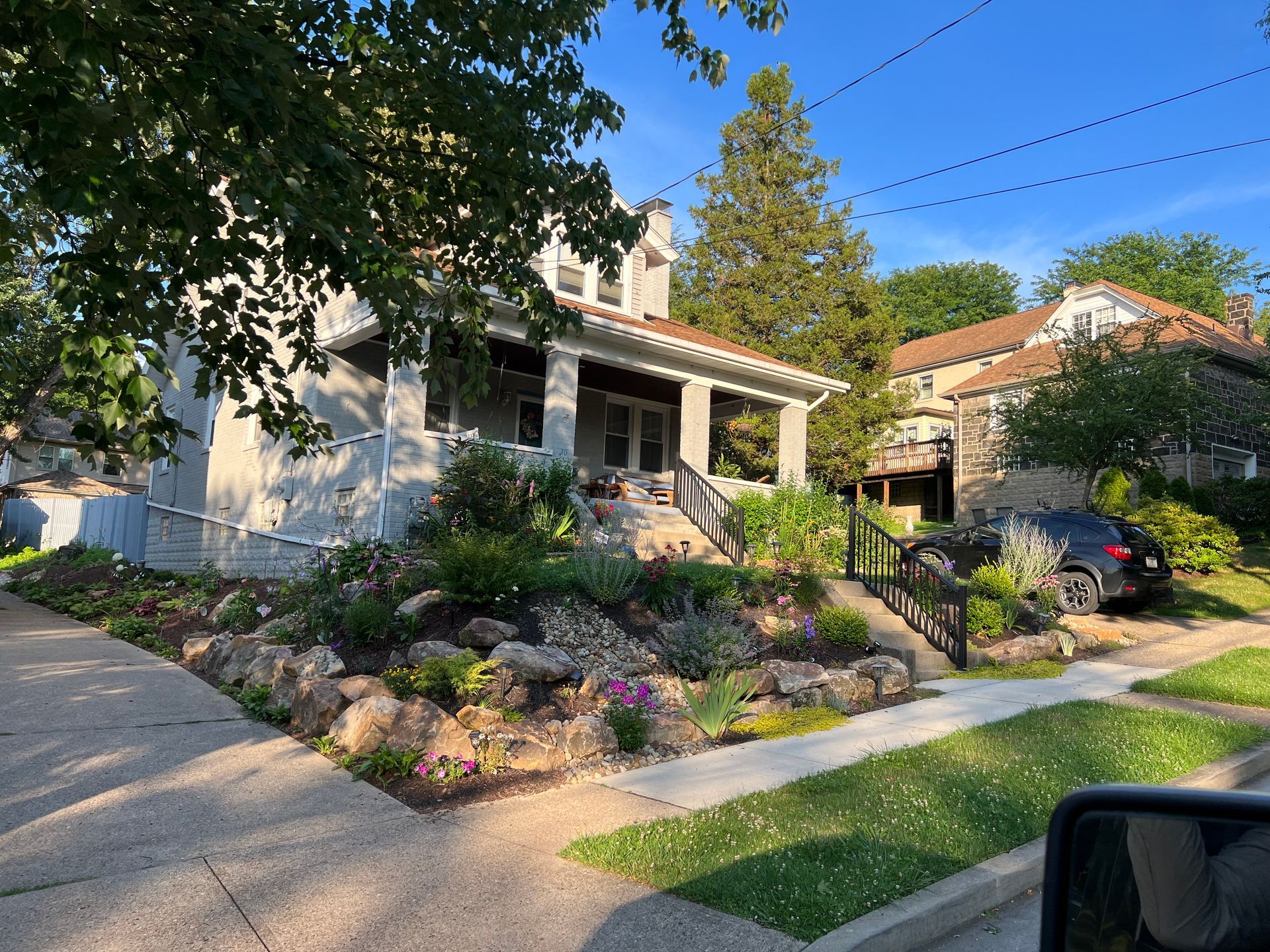 Sunny suburban house with a front porch, flower garden, driveway, and parked car