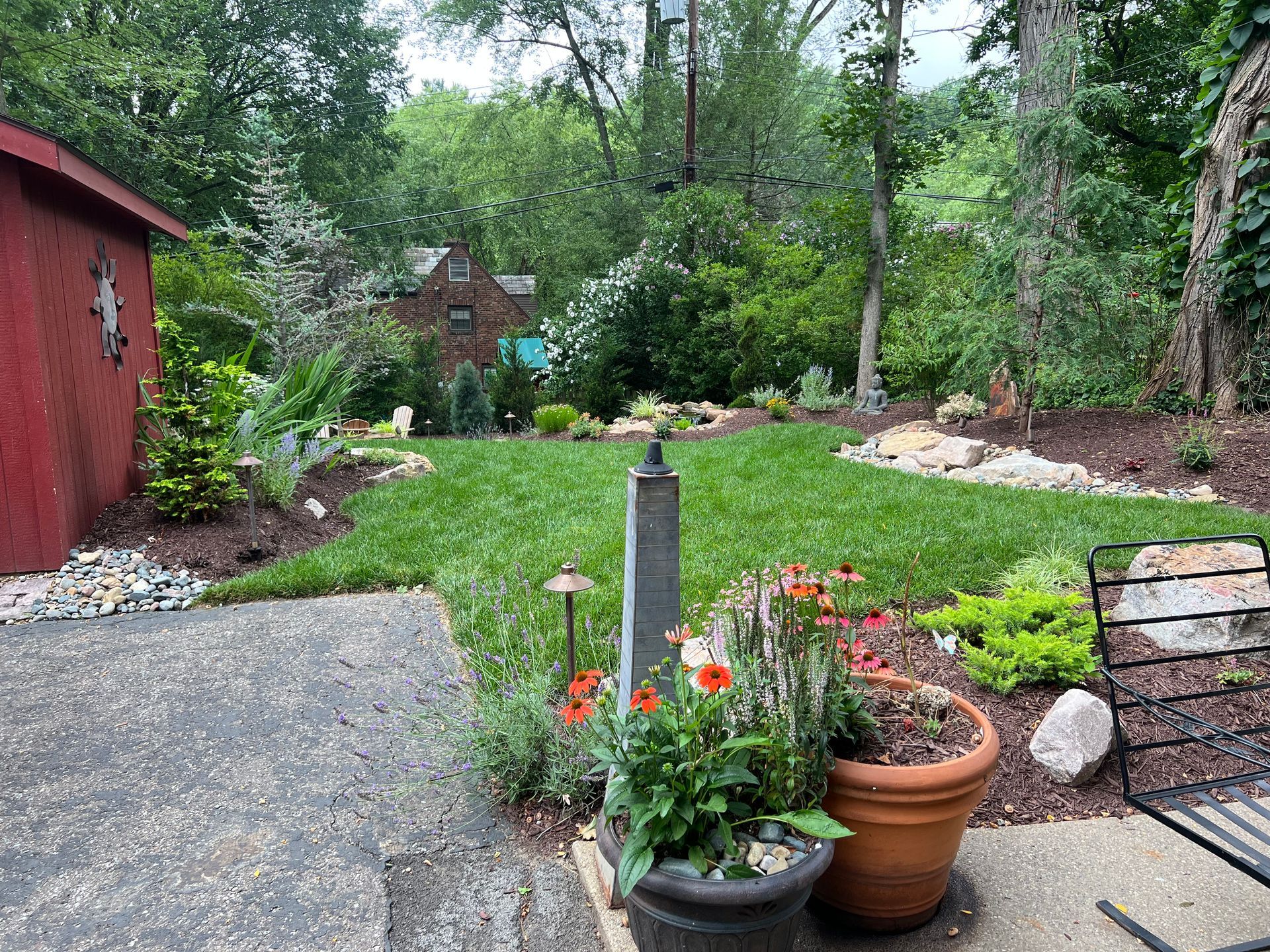 Garden path beside red shed, with potted flowers and a stone birdbath in a lush backyard.