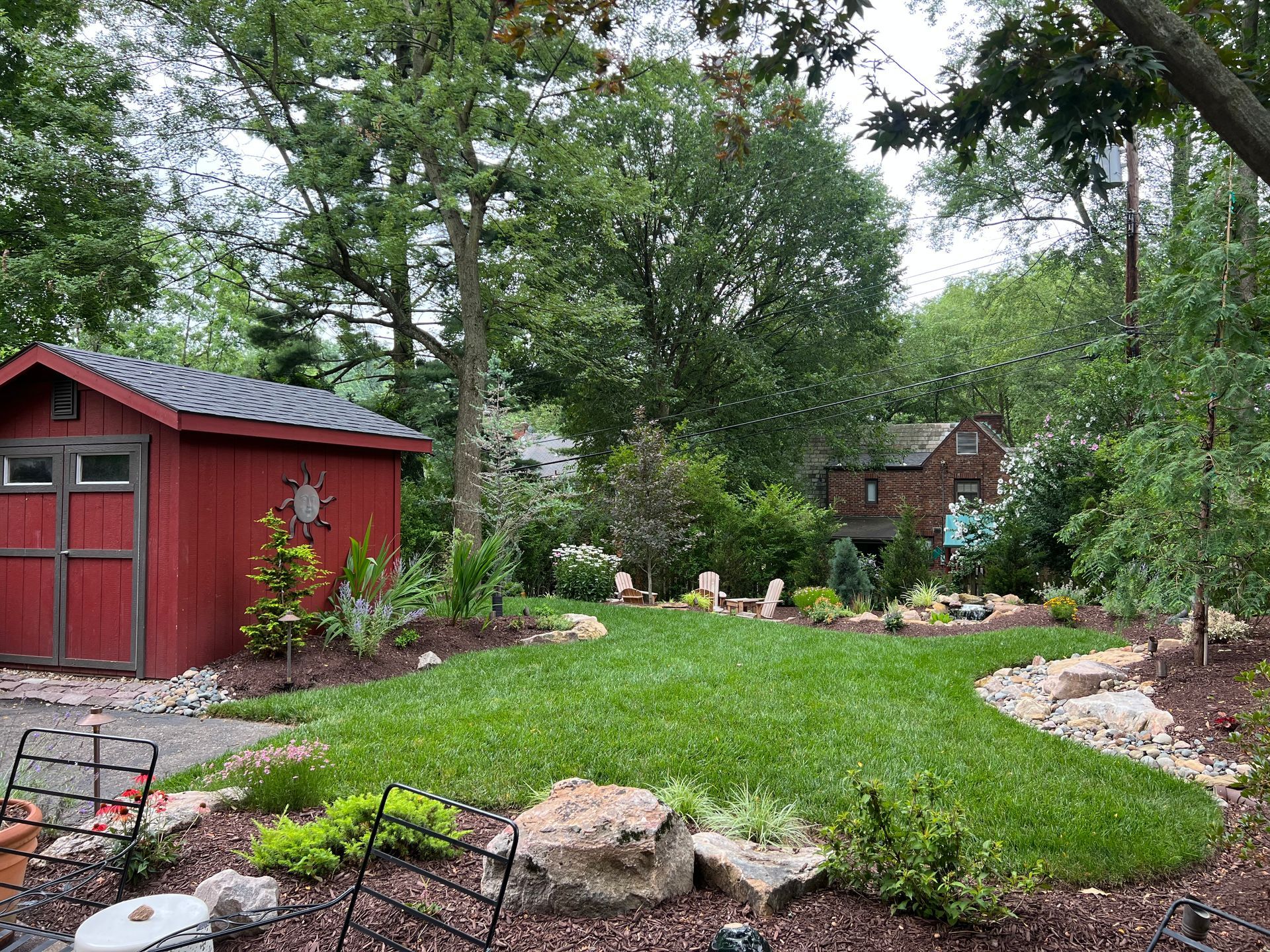 Red garden shed beside a green lawn with rocks, trees, and a flower bed in a wooded yard