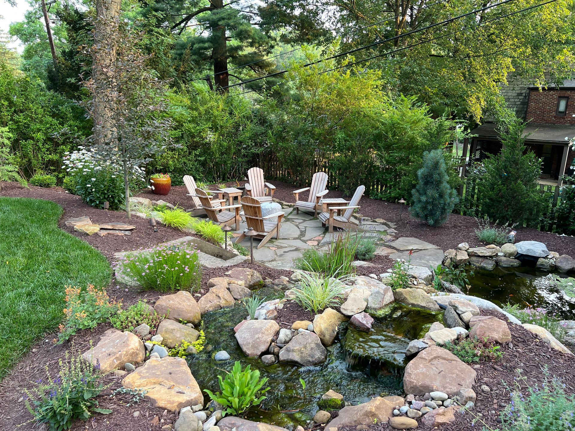Garden patio with stone path, white chairs, and lush greenery around a small rock pond