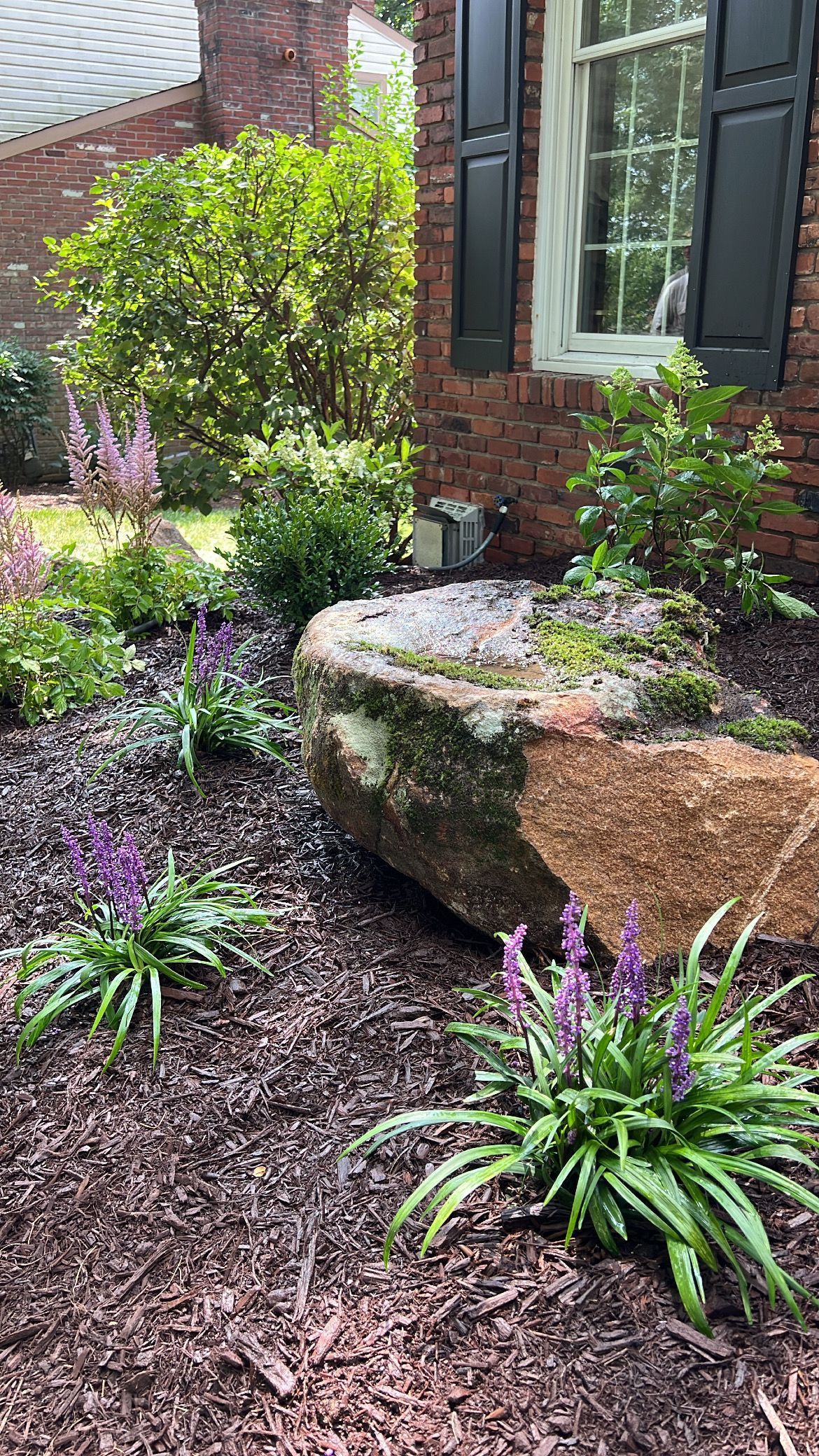 Garden bed with a mossy stone bench, purple flowers, and a brick house wall with shutters