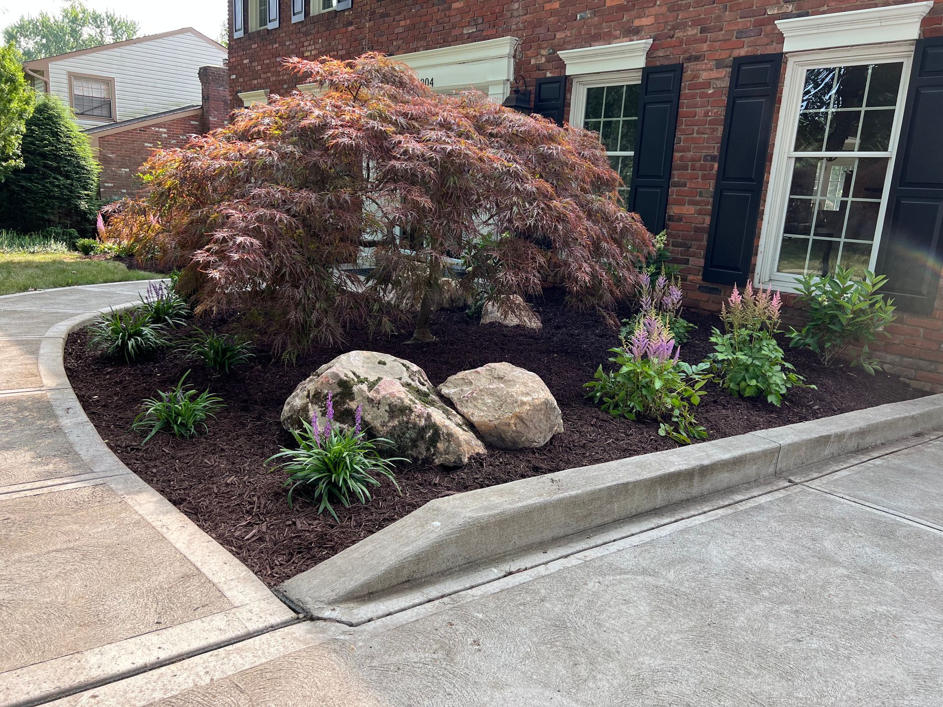 Landscaped corner with burgundy shrub, rocks, purple flowers, and a brick building beside a sidewalk