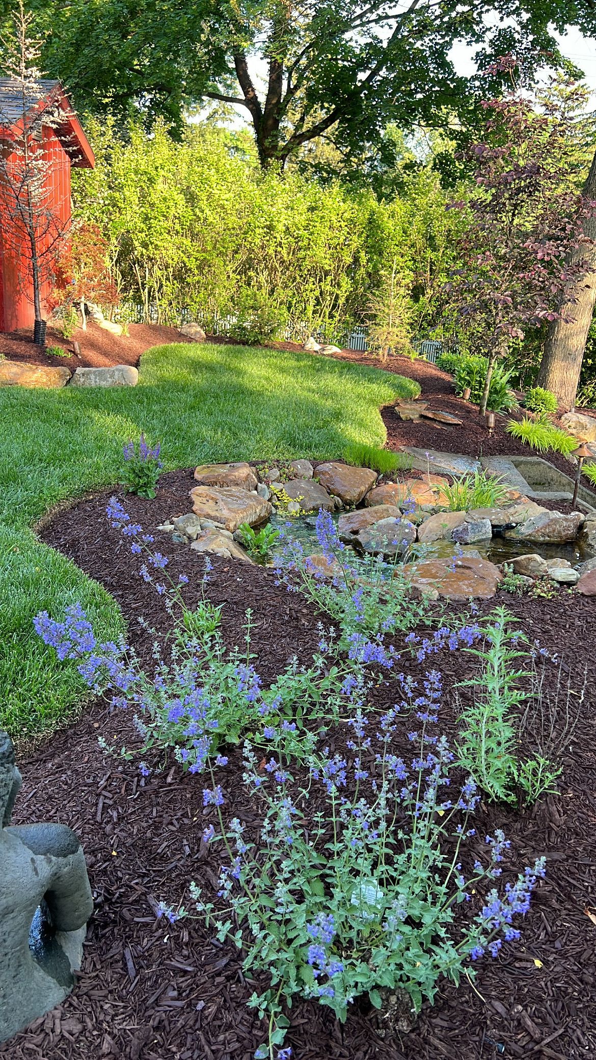 Sunlit garden with a stone path, green shrubs, and purple flowers beside a brick wall