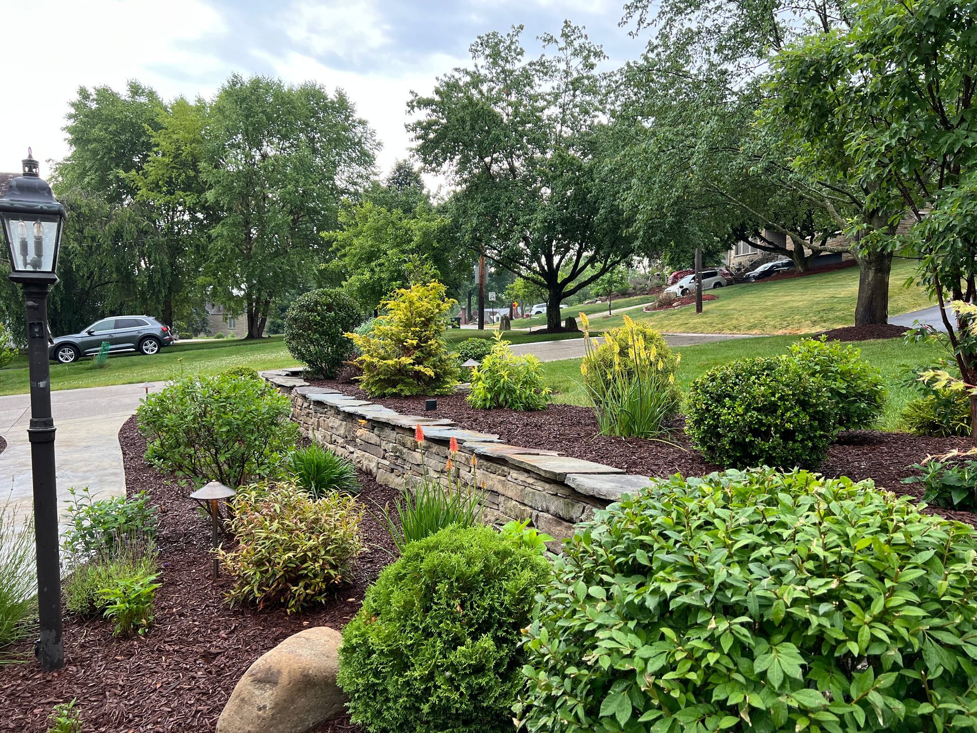 Landscaped garden with shrubs, stone wall, lamp post, and trees along a park path