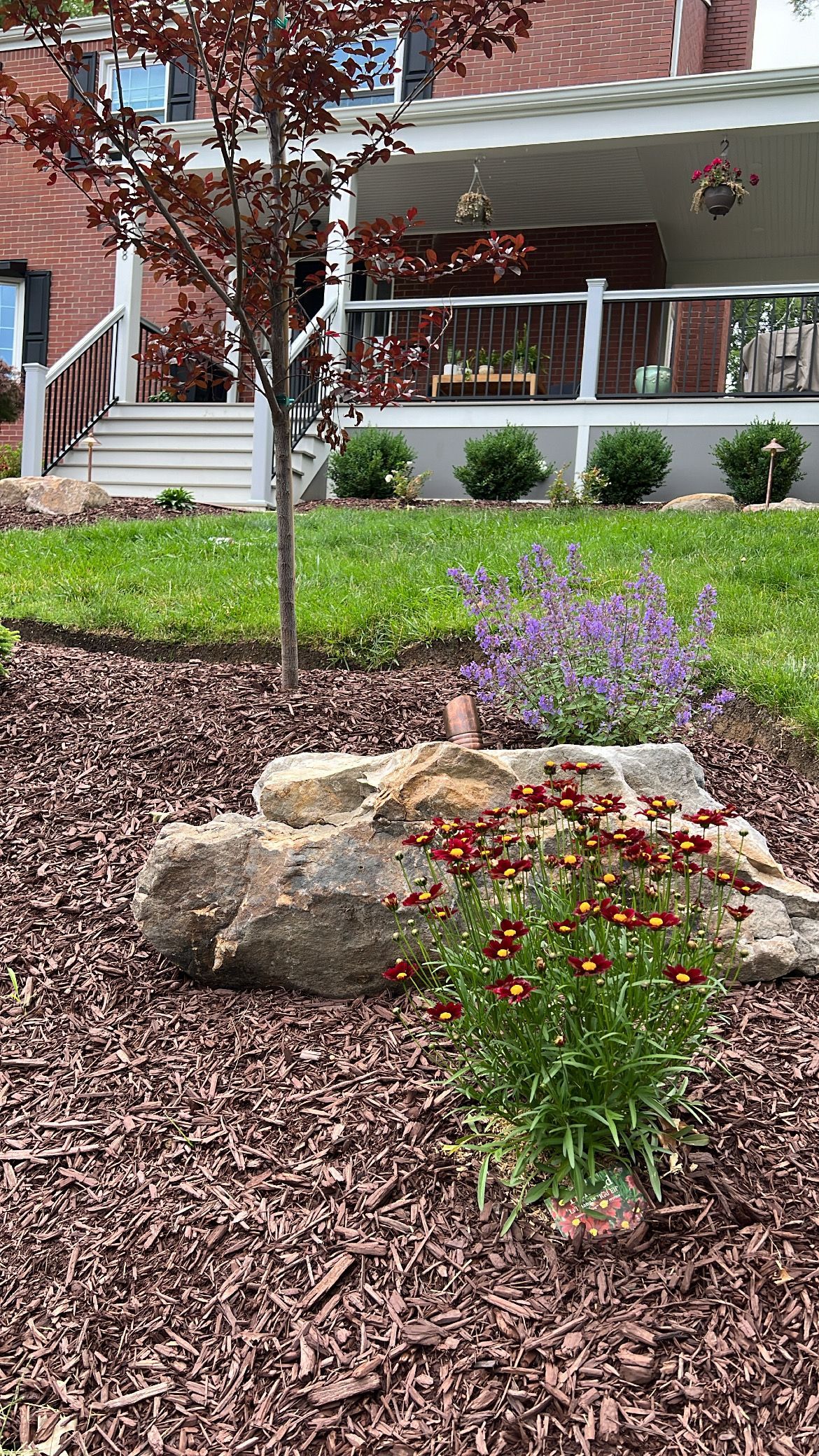 Flower bed with red and purple blooms, rocks, and mulch in front of a porch.