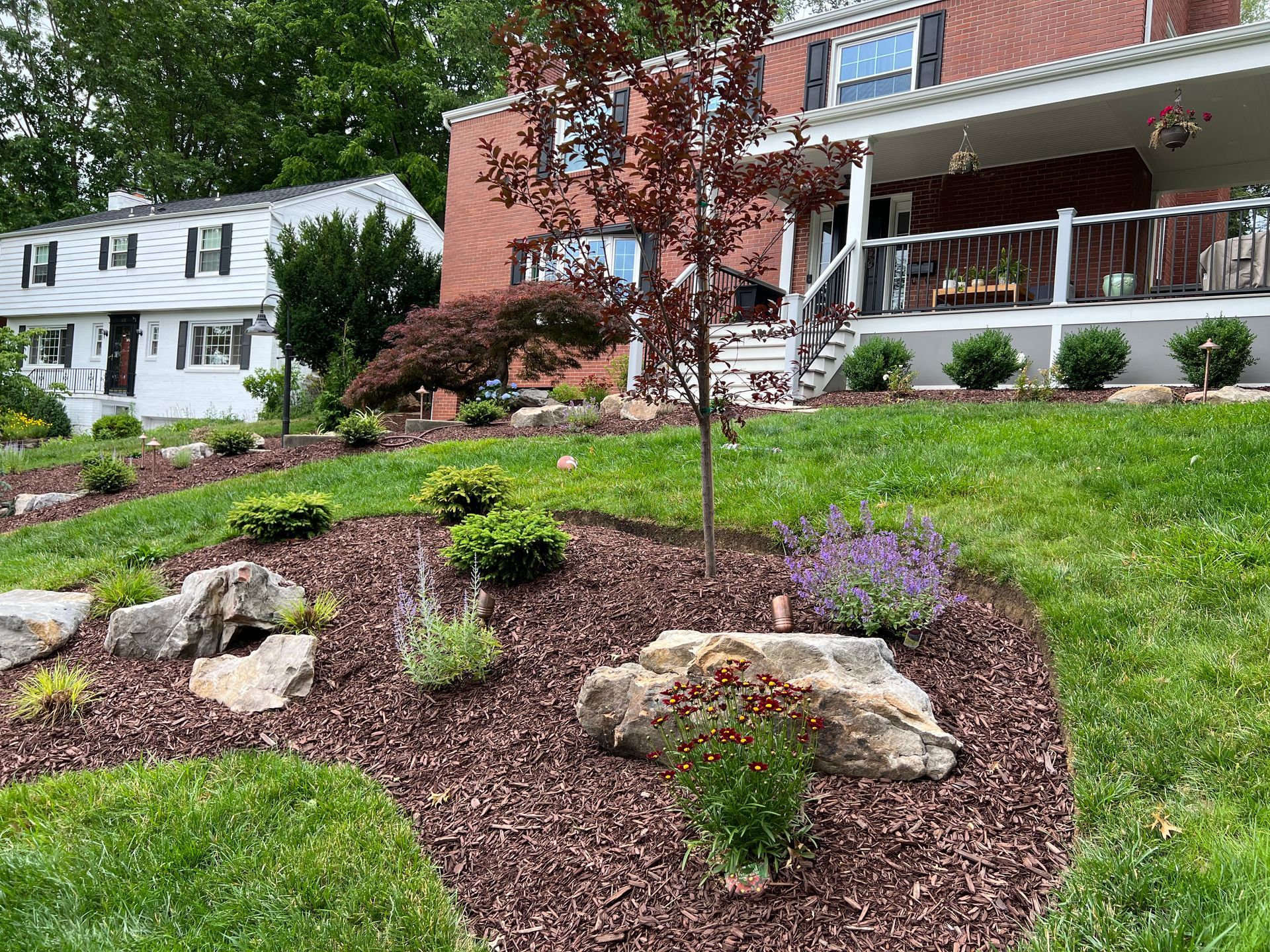 Landscaped front yard with mulch, rocks, shrubs, and a brick house with a porch in the background