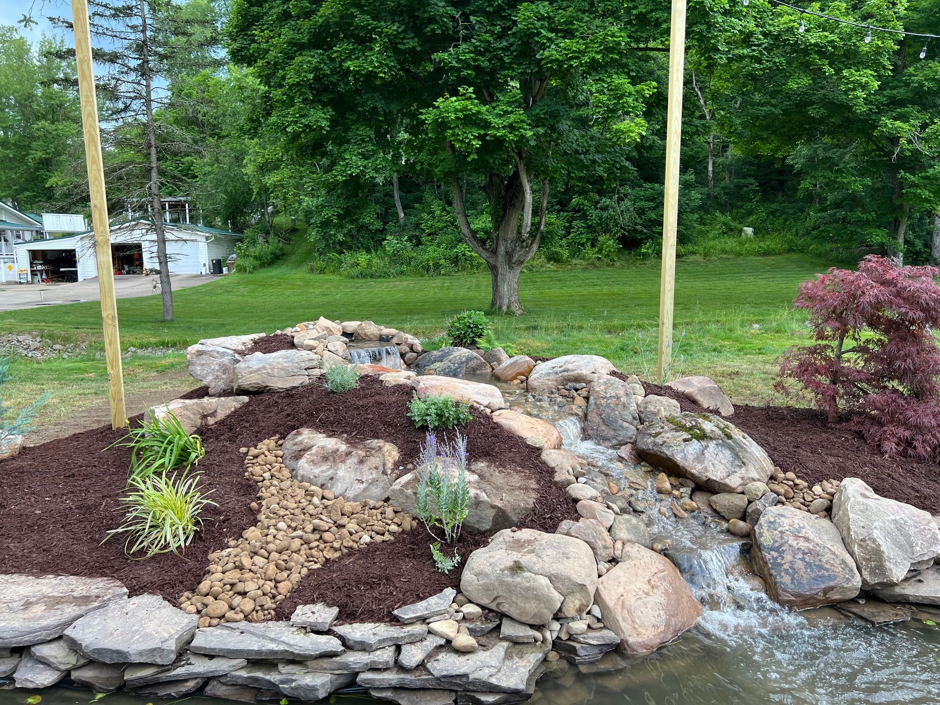 Rock garden with stacked stones, mulch, and a small waterfall beside a green lawn and trees
