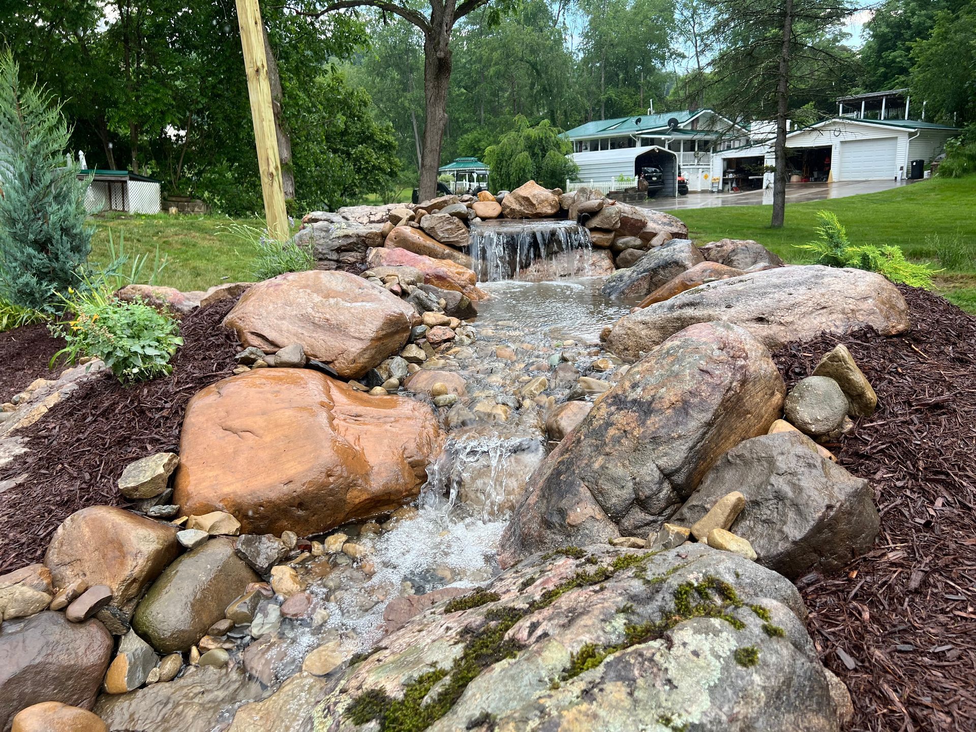 Rock garden stream waterfall with large tan boulders and trees in a suburban yard