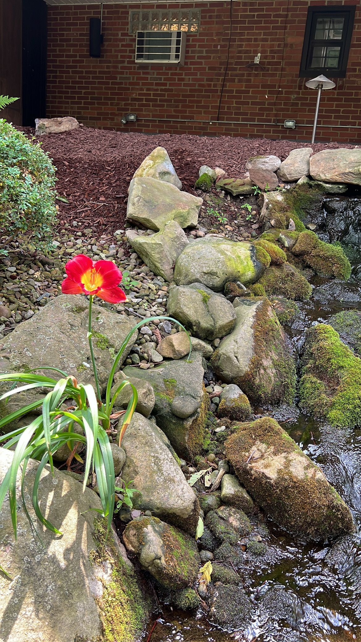 Garden waterfall over stacked rocks with moss and a bright red flower by a brick wall