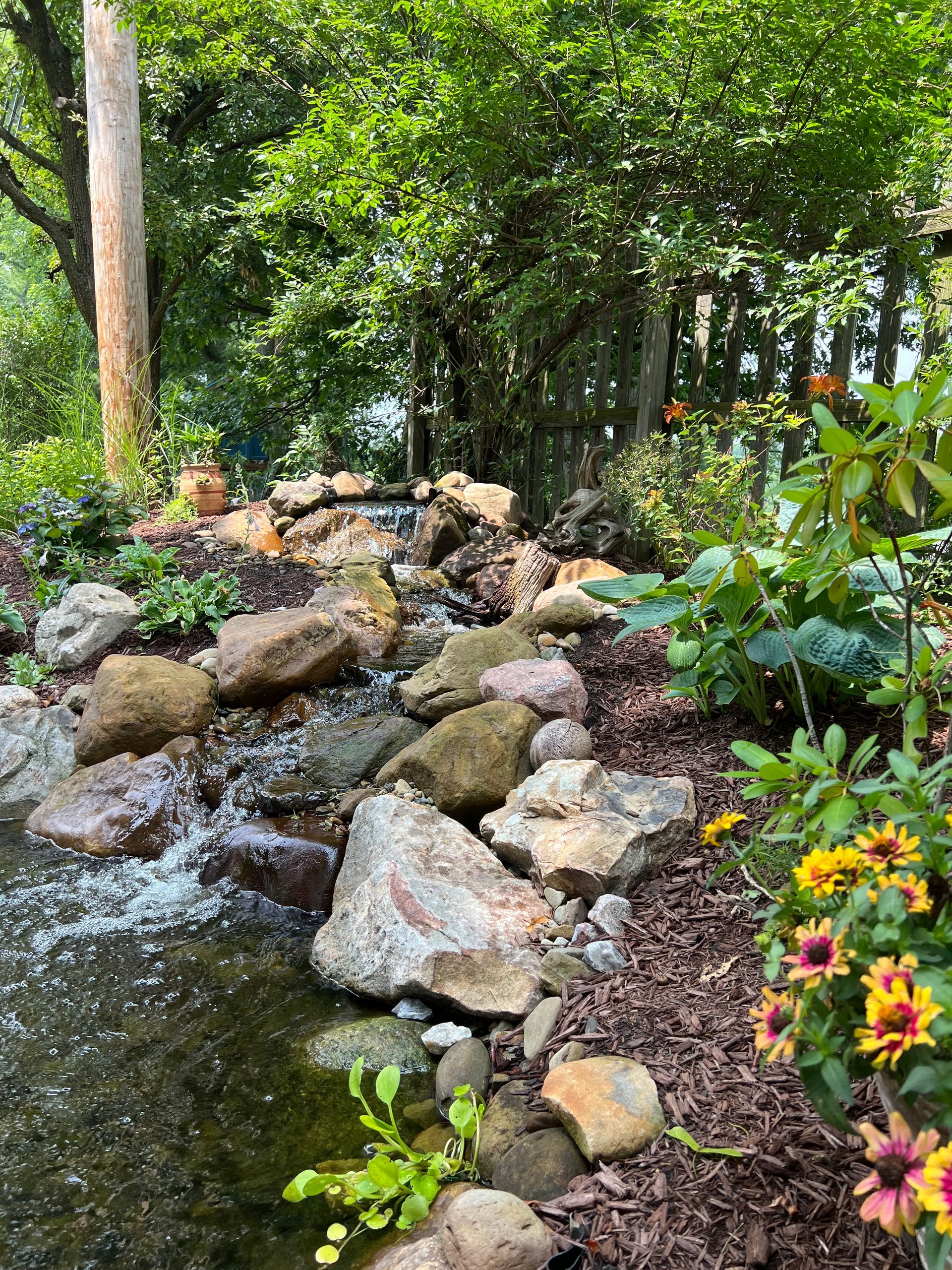 Rocky garden stream with flowing water, lush green trees, and colorful flowers along the bank