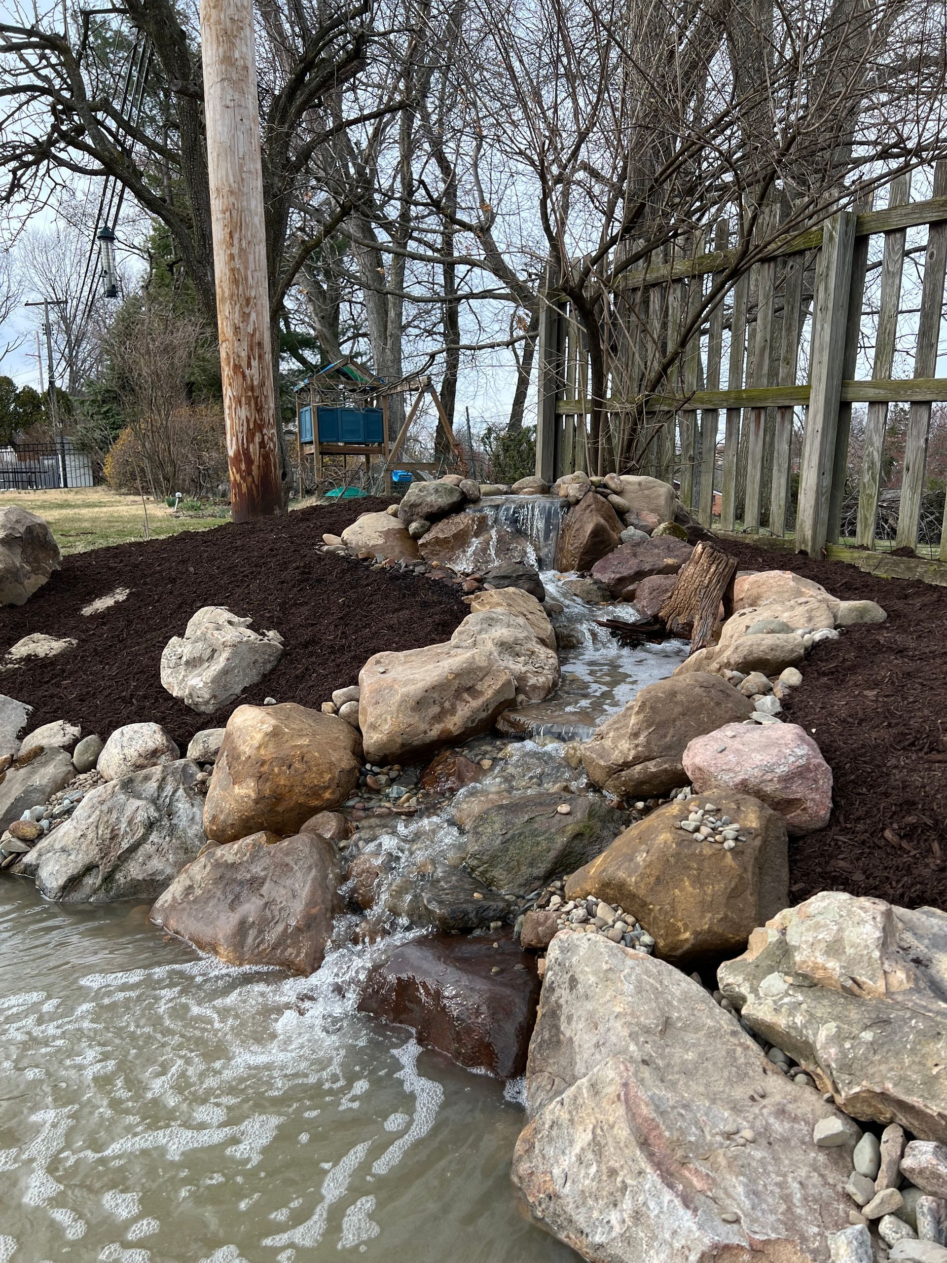 Rock-lined stream flowing through a wooded park beside leafless trees and a fence.